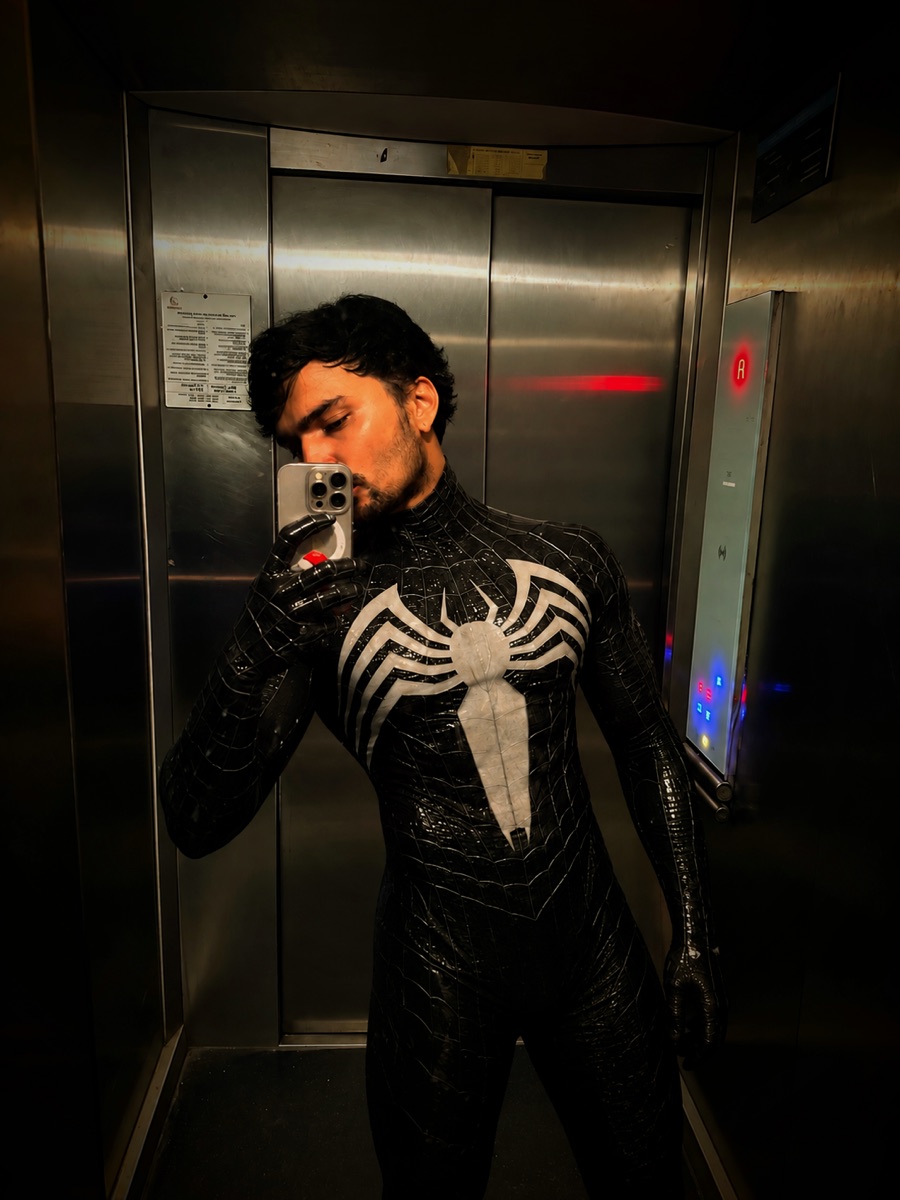 A man in a glossy black symbiote Spider-Man suit taking a mirror selfie in a steel elevator
